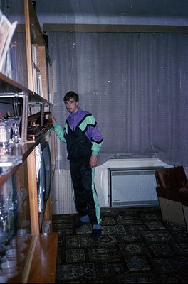 Vintage kitchen scene featuring a person in 1970s-style overalls (purple/blue/green) organizing glassware on a wooden shelf. ...