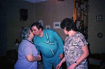 Vintage indoor scene featuring three adults in casual 1970s attire: a man in a teal track jacket, a woman in a patterned blou...