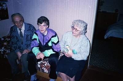 Three generations seated indoors, likely late 20th century. Elderly man in patterned sweater and tie, middle-aged woman in gl...