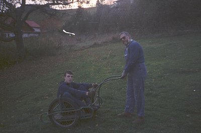 Two men in matching blue workwear assist a manual wheelchair in a rural setting, likely late 20th century. The standing man h...