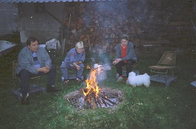 Three adults and a dog gather around an outdoor fire pit, cooking over flames. Mid-20th century clothing (striped shirts, car...