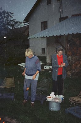 Two women in 1970s alpine retreat, preparing food under corrugated roof shelter. Left: woman in striped sweater and jeans hol...