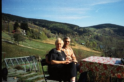 Two individuals pose outdoors on vintage wicker chairs beside a floral-patterned table, set against a lush, rolling countrysi...