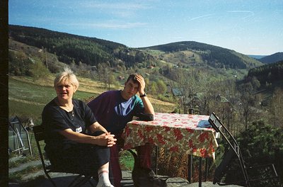 Two individuals pose outdoors at a rustic wooden table draped in floral-patterned red/white tablecloth, set on a concrete ter...