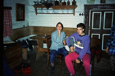 Vintage indoor scene featuring two individuals in a rustic, dimly lit room with wooden furniture. The girl wears a light blue...
