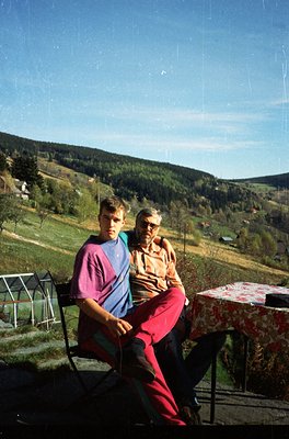 Vintage outdoor portrait of two individuals seated on wrought-iron chairs in a rural setting, likely Eastern Europe, 1970s. M...