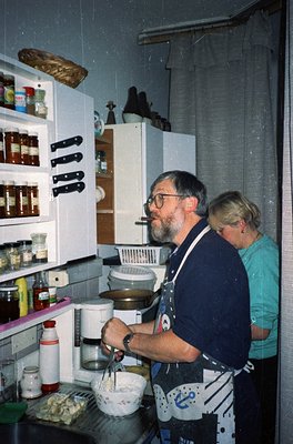 Vintage kitchen scene featuring two individuals preparing food in a modest, utilitarian space. The man in a patterned apron m...