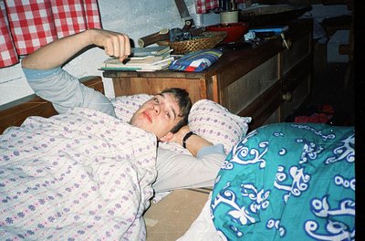 Vintage bedroom scene featuring a relaxed individual lying on floral-patterned bedding, holding a remote. Wooden furniture, i...