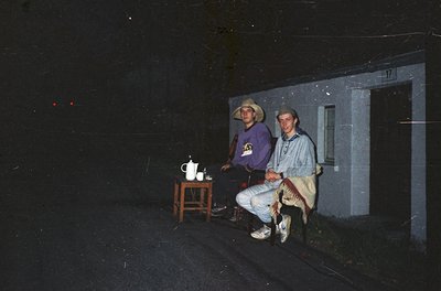 Vintage black-and-white photo of two men posing outside a plain concrete building. One wears a cowboy hat and a purple shirt ...