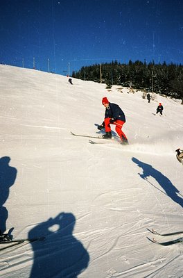 Vintage ski slope scene with skier in red cap and bright orange pants descending powdery snow. Other skiers visible in backgr...