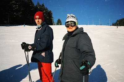 Two cross-country skiers pose on groomed trails in bright winter sunlight. The man wears a red knit cap, dark jacket, and red...