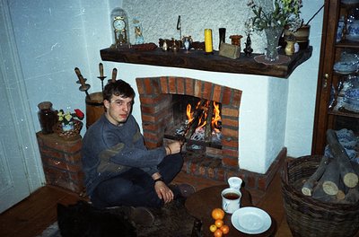 Vintage interior featuring a man seated by a brick fireplace with a warm fire. Surrounding decor includes brass candlesticks,...