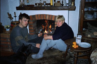 Two individuals seated by a brick fireplace in a rustic interior, likely Eastern European . The man wears a striped sweater a...