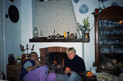 Two adults seated by a brick fireplace in a cozy, rustic interior. Wooden shelves display ceramic jars, blue-and-white patter...