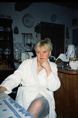 Vintage kitchen interior featuring a woman in a white robe, seated at a wooden table with blue-and-white patterned cloth. Sur...