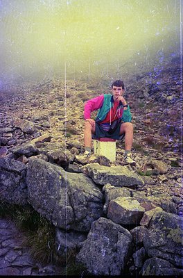 Hiker in vintage outdoor gear poses on rocky alpine trail, misty backdrop suggests high elevation. Brightly colored jacket an...