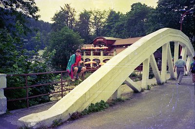 Vintage wooden bridge slide at a lakeside resort, likely from the 1960s–70s. Two children descend a curved slide into a shall...
