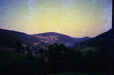 Vintage slide scan of a mountainous valley bathed in golden evening light, with scattered streaks from film damage. Rural set...