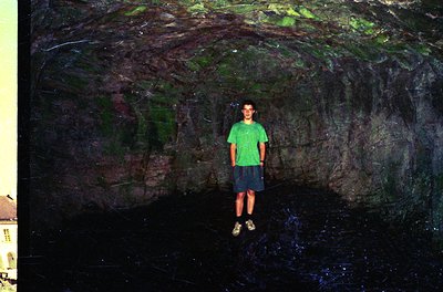 A young individual stands in a narrow, moss-covered rock crevice with water at the base, likely a natural cave or grotto. The...