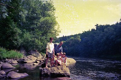 Three individuals pose on river rocks during golden-hour lighting, mid-1970s. Woman in patterned skirt and blouse stands besi...