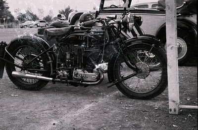 Vintage motorcycle with sidecar frame, likely a pre-1940s AJ model, parked outdoors near classic cars. Chrome engine and dual...