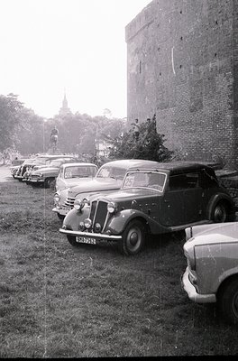 Classic 1940s–1950s vintage cars parked beside a brick fortress wall, likely European. Distinctive rounded fenders and chrome...