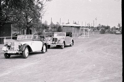 Vintage roadside scene featuring three classic convertibles parked in a row, likely mid-20th century (1940s–1950s). Distincti...