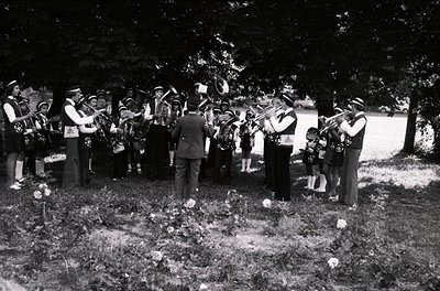 A black-and-white photograph captures a solemn outdoor funeral procession, likely from the **1950s–1960s**. A brass band in f...