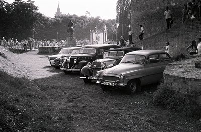 Classic 1960s European rally scene: vintage cars (Volvo PV544 & Fiat 1200) parked on gravel beside a stone-walled hillside. C...