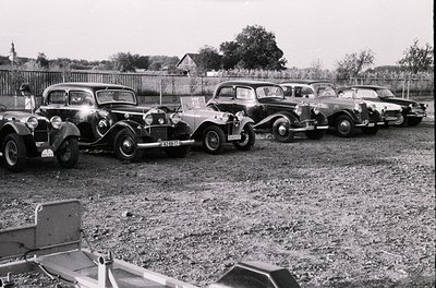 Six vintage cars lined up in a gravel lot, likely mid-20th century (1940s–1960s). Distinctive models include a pre-war conver...