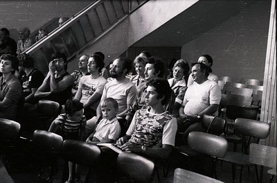 A black-and-white photo of a mixed-gender audience seated in tiered theater-style seating, likely from the 1970s–1980s. Casua...