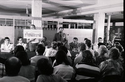 Black-and-white photo of a mid-20th-century lecture or meeting in an industrial hall. A speaker at a podium holds a poster wi...