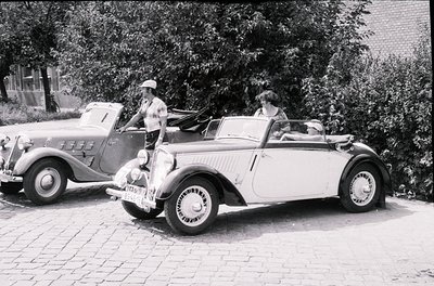 Vintage 1930s–1940s roadside scene featuring a white convertible with streamlined bodywork and wire-spoke wheels, parked besi...