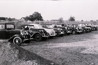 Classic vintage cars lined up in a gravel lot, likely a 1930s–1940s European setting. Distinctive pre-war designs, including ...