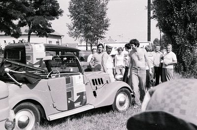 Vintage rally car with sponsor decals and mud splatter, surrounded by a group of casually dressed spectators in 1970s outdoor...
