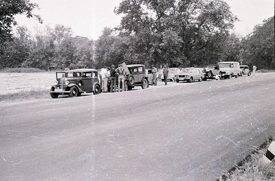 Vintage roadside gathering of vintage cars (1930s–1940s) lined up on a rural road, surrounded by dense trees. Group of people...