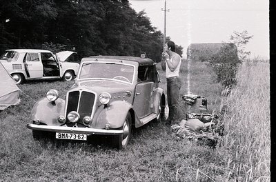 Vintage 1950s-era sedan (BMA-73-62) parked in grassy field with classic rounded headlights and chrome accents. Man in white s...