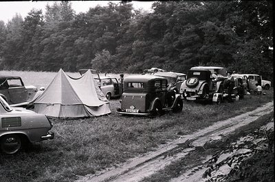 Vintage camping scene with classic cars and tents along a lakeside dirt road, mid-20th century. Vehicles include a vintage tr...