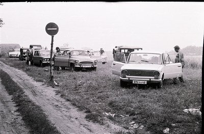 Vintage black-and-white roadside scene with 1970s-era cars parked on grass beside a rural road. Soviet-era vehicles dominate,...