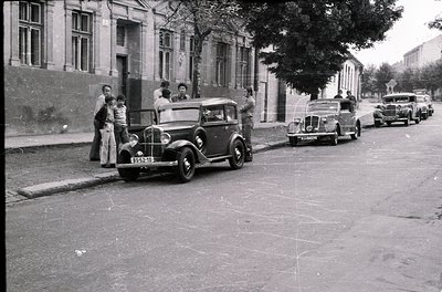 Vintage black-and-white street scene featuring 1930s-era cars parked along a cobblestone road. Group of people in early 20th-...