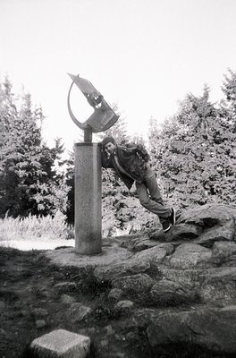 Black-and-white photo of a person adjusting a large, abstract sundial sculpture atop a stone pedestal in a forested area. The...