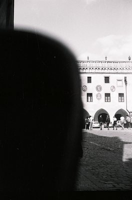 Black-and-white shot of a cobblestone plaza framed by a blurred silhouette. Prominent neoclassical building with arched entra...