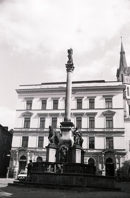Neoclassical monument with tall column and sculptural reliefs in front of a multi-story institutional building, likely from t...
