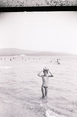 Mid-20th century black-and-white seaside scene: boy in swim trunks stands waist-deep in shallow water, adjusting a hat. Crowd...
