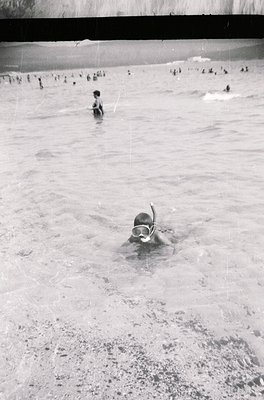 Black-and-white shot of a crowded outdoor swimming pool, likely from the 1950s–1970s. A child in a snorkel mask dives near sh...