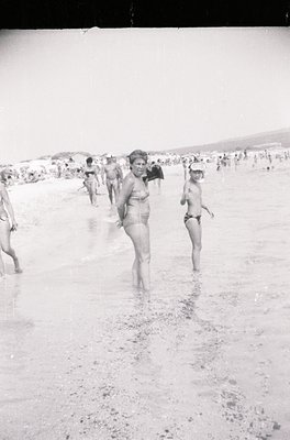 Black-and-white beach scene from the 1960s–70s, featuring mid-century swimwear and relaxed coastal culture. Central figure in...