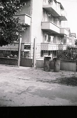 Mid-century urban courtyard with Soviet-era apartment block. Concrete balconies, barred windows, and a gated entrance sign hi...