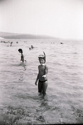 Black-and-white beach scene from mid-20th century, likely 1950s–1960s. A young boy in swim trunks and snorkel stands in shall...