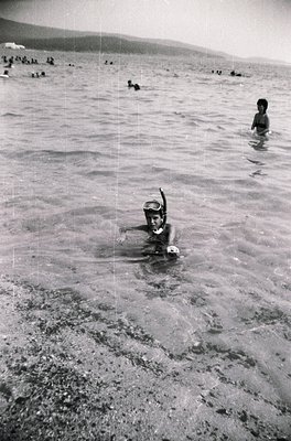 Black-and-white snapshot of a mid-20th century seaside scene. A child snorkels in shallow water, wearing a snorkel mask and s...