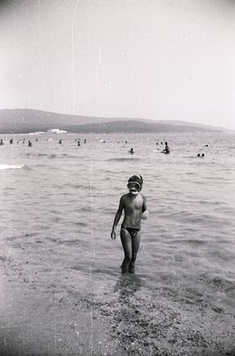 Black-and-white beach scene from the 1960s–70s, featuring a child in swim trunks and goggles wading into shallow water. Crowd...